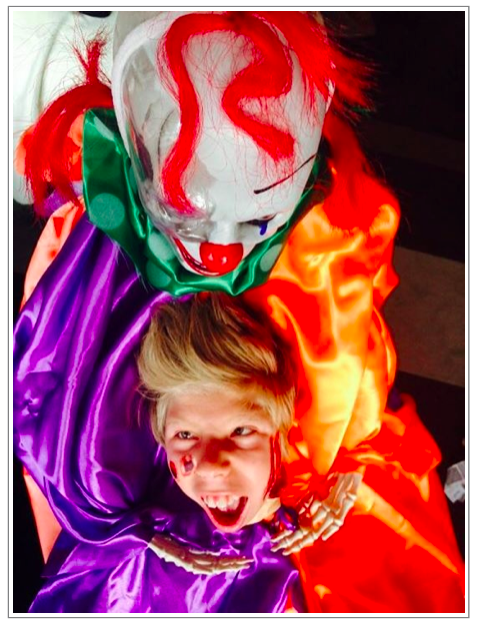 looking down from above the skeleton clown head with red flashes of hair, shiny purple and orange satin shirt and one very happy boy with just his head exposed as though the clown is holding his severed head - halloween creeps.